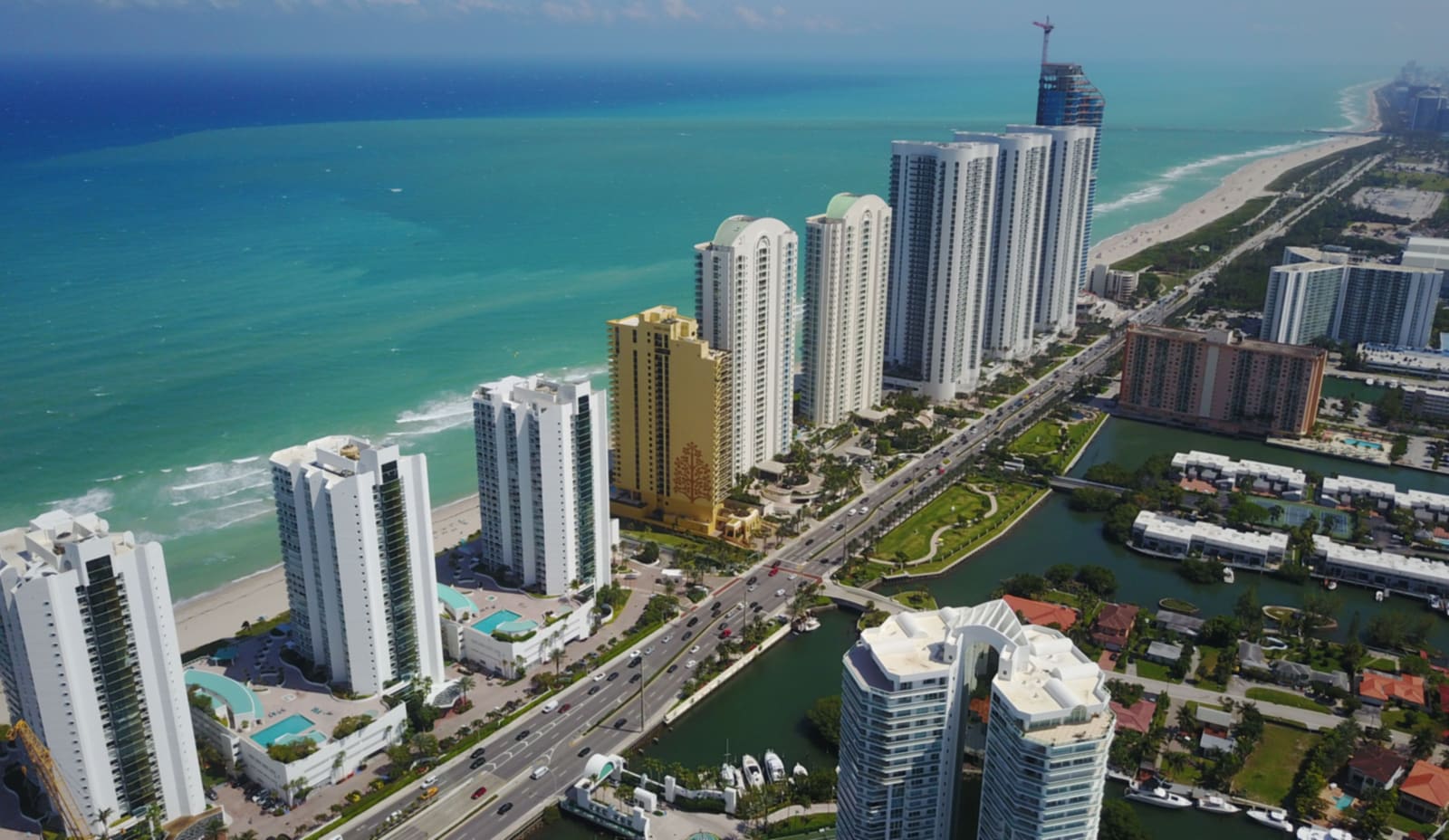 Aerial view north along Sunny Isles Beach: a line of oceanfront condominium towers fronting the Atlantic, with Collins Avenue and the Intracoastal waterway behind them